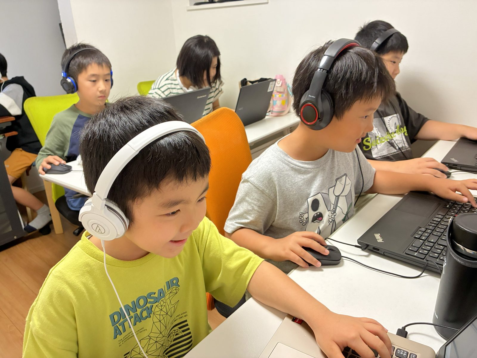 ヘッドホンをつけた子どもたちが、笑顔でそれぞれのパソコンに向かってプログラミングに取り組む教室の様子